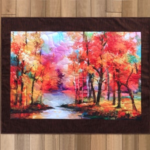 Puede incluir: Un tapiz de pared acolchado que presenta una vibrante escena de bosque otoñal. La obra de arte muestra árboles con follaje rojo, naranja y amarillo, un arroyo y un cielo colorido. La pieza está enmarcada con un borde marrón oscuro y se exhibe sobre un fondo de madera.