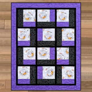 Op de afbeelding: Een rechthoekige quilt met een paarse rand en een raster van twaalf vierkante blokken. Elk blok toont een witte achtergrond met een cirkelvormige afbeelding van een pompoen, een heksenhoed en de tekst Happy Halloween, omringd door zwarte en paarse stof.