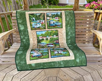 May include: A quilted throw blanket with four rectangular panels depicting vintage caravans in outdoor scenes. The blanket has a green border and a cream centre, displayed on a wooden swing.
