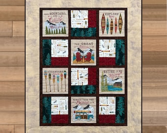 May include: A quilted wall hanging with nine square panels. Each panel features outdoor-themed imagery and text, including mountains, canoes, and phrases like "Adventure Awaits" and "Explore More." The colour palette includes shades of brown, red, green, and cream.
