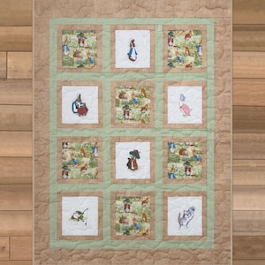 May include: A rectangular quilt featuring a grid of twelve square panels. The panels alternate between white fabric with embroidered Peter Rabbit characters and green fabric with a repeating Peter Rabbit print. The quilt has a tan border.