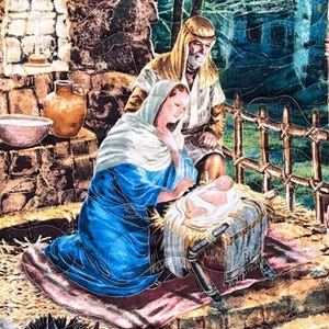 Könnte beinhalten: Eine gesteppte Darstellung der Weihnachtsgeschichte. Maria, in einem blauen Gewand und mit weißer Kopfbedeckung, kniet neben dem Jesuskind in einer Krippe. Josef steht hinter ihnen. Die Szene spielt in einem rustikalen Stall.