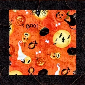 Op de afbeelding: Een vierkant stoffen quiltblok met een centraal oranje paneel met een Halloween-print van geesten, pompoenen, uilen, zwarte katten, ketels en vleermuizen, inclusief de tekst BOO, omringd door een zwarte rand met een gestikt wervelpatroon.