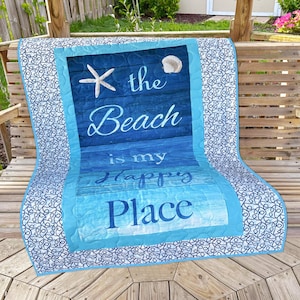 Può includere: Una trapunta blu e bianca con un design a tema spiaggia. La trapunta presenta una stella marina, una conchiglia e il testo "The Beach is my Happy Place".