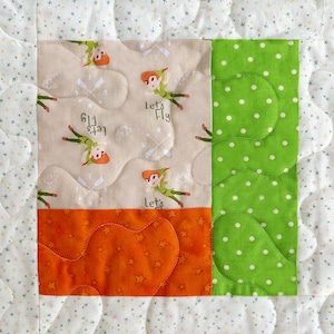 May include: A quilted fabric square featuring a whimsical design. The square is divided into sections with different patterns: a tan section with Peter Pan illustrations and the text "Let's Fly", a green polka-dot section, and an orange section with star shapes.