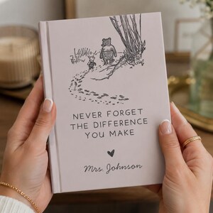 Puede incluir: Un cuaderno de tapa dura gris claro con un dibujo lineal negro de Winnie the Pooh y Piglet caminando por un sendero. El texto dice "NEVER FORGET THE DIFFERENCE YOU MAKE", seguido de un pequeño corazón y el nombre "Mrs. Johnson".