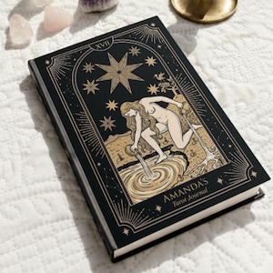 Può includere: Un diario di tarocchi nero con illustrazioni dorate e la scritta "AMANDA'S Tarot Journal". La copertina presenta un design celeste con una stella e una figura che versa acqua. Cristalli e un candelabro sullo sfondo.