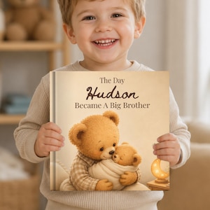 Könnte beinhalten: Ein gebundenes Buch mit dem Titel The Day Hudson Became A Big Brother. Das Cover zeigt eine Illustration eines großen Teddybären, der einen kleineren, eingewickelten Teddybären hält. Eine kleine, leuchtende Mondsichel-Lampe steht auf einem Tisch.