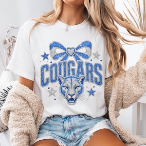 May include: A white short-sleeve t-shirt featuring a graphic print of a blue glittery bow, a heart, and a cougar face. The text COUGARS is printed in large blue glittery letters. The design includes small blue and silver glittery star accents.