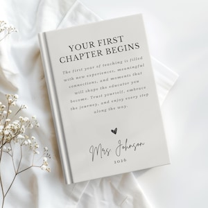 May include: A light gray hardcover book with the text "YOUR FIRST CHAPTER BEGINS" followed by a paragraph about the first year of teaching, a small heart icon, the name "Mrs. Johnson," and the year "2026" printed in black ink on the front cover.