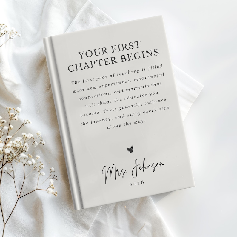 May include: A light gray hardcover book with the text "YOUR FIRST CHAPTER BEGINS" followed by a paragraph about the first year of teaching, a small heart icon, the name "Mrs. Johnson," and the year "2026" printed in black ink on the front cover.