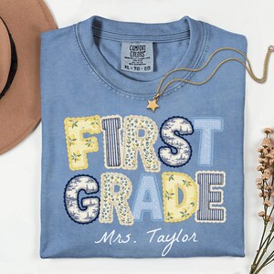 以下が含まれることがあります： FIRST GRADEとMrs. Taylorの白い文字がプリントされた青い半袖Tシャツ。文字の中には、黄色いレモン、青いストライプ、花柄など、さまざまな模様が描かれています。襟元にはゴールドの星のネックレスが置かれています。