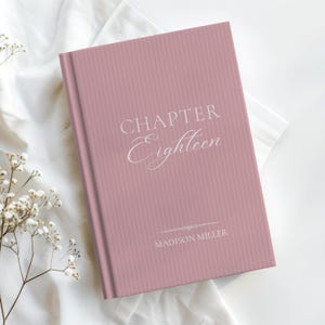 Pode incluir: Um livro de capa dura rosa empoeirado com um padrão de riscas verticais. A capa apresenta as palavras "CHAPTER Eighteen" em escrita delicada, com "MADISON MILLER" por baixo. O livro repousa sobre um tecido branco, com raminhos de flores brancas.
