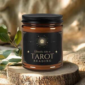 May include: A brown candle in a glass jar with a black lid. The label reads "Smells like a Tarot Reading" with a sun and moon design. The candle sits on a wooden slice, with greenery in the background.