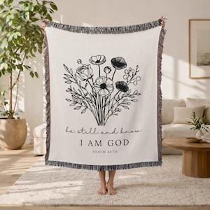 May include: Cream-coloured woven throw blanket featuring a black floral design and the text "be still and know I AM GOD" from Psalm 46:10. The blanket has a fringed edge and is displayed indoors.