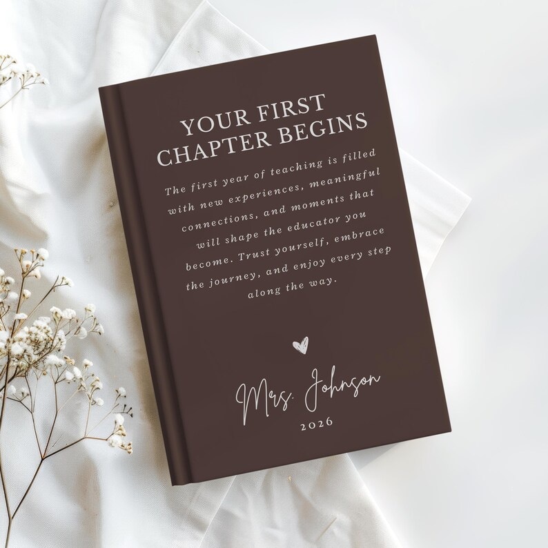 May include: A dark brown hardcover book with white text that reads: YOUR FIRST CHAPTER BEGINS. The first year of teaching is filled with new experiences, meaningful connections, and moments that will shape the educator you become. Mrs. Johnson 2026.