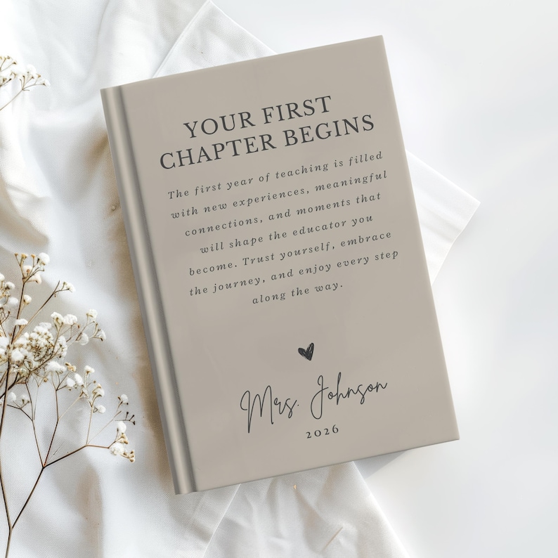 May include: A light beige hardcover book with the text "YOUR FIRST CHAPTER BEGINS" followed by a paragraph about the first year of teaching, a small heart icon, the name "Mrs. Johnson," and the year "2026" printed in dark gray ink on the cover.