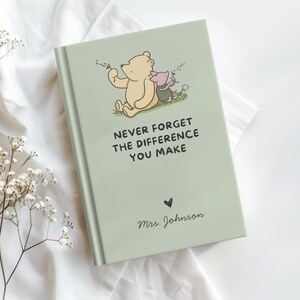 May include: A light sage green hardcover book featuring a printed illustration of Winnie the Pooh and Piglet sitting together. The text reads, "NEVER FORGET THE DIFFERENCE YOU MAKE" above a small heart and the name "Mrs. Johnson" in script.