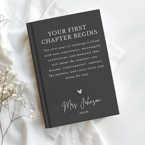 May include: A dark gray hardcover book with white text that reads: YOUR FIRST CHAPTER BEGINS. The first year of teaching is filled with new experiences, meaningful connections, and moments that shape the educator you become. Mrs. Johnson 2026.