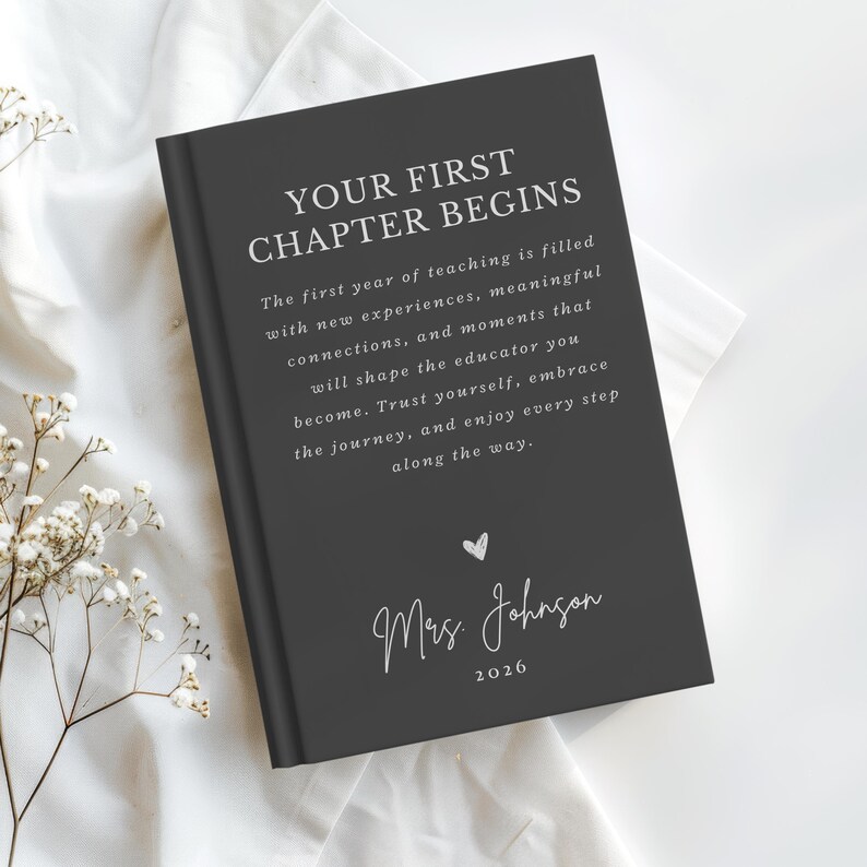 May include: A dark gray hardcover book with white text that reads: YOUR FIRST CHAPTER BEGINS. The first year of teaching is filled with new experiences, meaningful connections, and moments that shape the educator you become. Mrs. Johnson 2026.