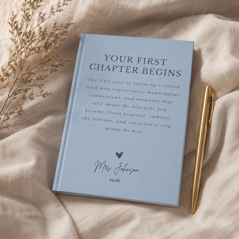 May include: A light blue hardcover journal with the text "YOUR FIRST CHAPTER BEGINS" followed by an inspirational paragraph, a small heart icon, the name "Mrs Johnson", and the year "2026" printed in dark blue, accompanied by a gold pen.