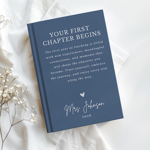 May include: A navy blue hardcover book with white text that reads: YOUR FIRST CHAPTER BEGINS. The first year of teaching is filled with new experiences, meaningful connections, and moments that will shape the educator you become. Trust yourself, embrace the journey, and enjoy every step along the way. Below is a small heart icon, the name Mrs. Johnson, and the year 2026.