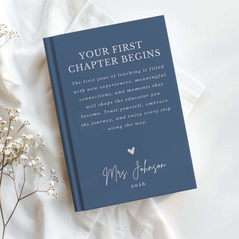 May include: A navy blue hardcover book with white text that reads: YOUR FIRST CHAPTER BEGINS. The first year of teaching is filled with new experiences, meaningful connections, and moments that will shape the educator you become. Trust yourself, embrace the journey, and enjoy every step along the way. Below is a small heart icon, the name Mrs. Johnson, and the year 2026.