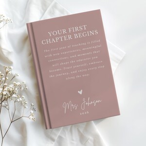 May include: A dusty rose hardcover book with white text that reads: YOUR FIRST CHAPTER BEGINS. The first year of teaching is filled with new experiences, meaningful connections, and moments that will shape the educator you become. Mrs. Johnson 2026.