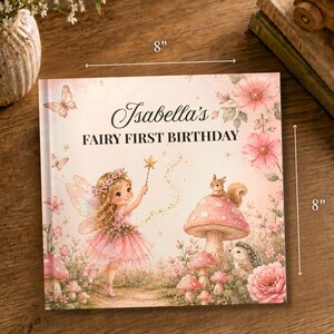 May include: A square book titled Isabella's FAIRY FIRST BIRTHDAY, measuring 8 inches by 8 inches. The cover features a fairy with wings, a wand, and a pink dress, surrounded by mushrooms, flowers, a squirrel, and a hedgehog in soft pink tones.