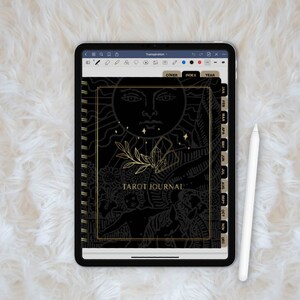 May include: A black and gold tarot journal with a spiral binding. The cover features a sun with a face and a crystal design. The journal has tabs for each month of the year.