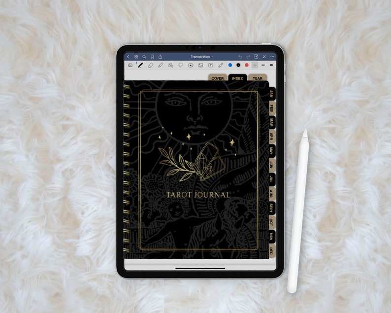 May include: A black and gold tarot journal with a spiral binding. The cover features a sun with a face and a crystal design. The journal has tabs for each month of the year.
