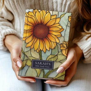 May include: A notebook for Bible study with a stained-glass effect sunflower design. The cover is personalised with the name "Sarah's" and the text "Bible Study Notes" on a rectangular label. The sunflower has yellow petals and a brown centre.