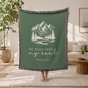 May include: Green woven throw blanket featuring a white mountain and river landscape design. The text "HE RESTORES my soul" and "PSALM 23:3" are printed on the blanket. The blanket has a fringed edge.