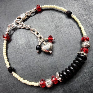 May include: A beaded bracelet featuring cream-colored seed beads, faceted black and red glass beads, and silver-toned metal accents. The bracelet includes a silver-toned heart charm dangling from a chain link extension with a lobster claw clasp.