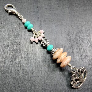 May include: A decorative charm featuring a silver lobster clasp, turquoise and light pink beads, silver-toned metal spacers, three speckled tan oval beads, and a silver-toned lotus flower pendant at the base, all connected by a silver wire.