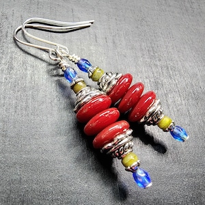 May include: A pair of dangle earrings featuring silver-tone hooks, each adorned with a stack of three flat, circular red beads, interspersed with textured silver-tone spacers, and finished with small, round yellow and faceted blue glass beads.