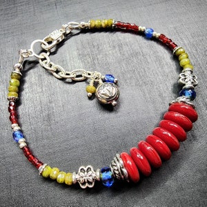 May include: A beaded bracelet featuring a series of large, flat, circular red beads, smaller red and olive green seed beads, and vibrant blue crystal accents. The design includes silver-toned metal spacers, a lobster clasp, and a dangling charm.