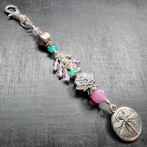 May include: A decorative charm featuring a silver lobster clasp, a metallic bead, turquoise and light pink faceted beads, a silver lotus flower charm, a smooth round pink bead, and a circular silver pendant embossed with a dragonfly design.
