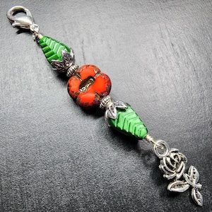 May include: A decorative charm featuring a silver-tone lobster clasp, two green leaf-shaped glass beads with silver-tone metal caps, a central orange flower-shaped glass bead, and a silver-tone metal rose charm at the bottom end of the piece.