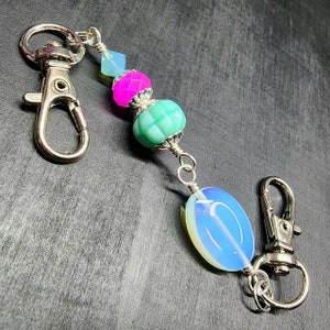 Peut inclure: Une chaîne décorative composée de deux fermoirs mousquetons en métal argenté reliés par une série de perles. Les perles incluent une petite perle à facettes bleu clair, une perle à facettes rose vif, une perle verte texturée et une grande perle ovale.