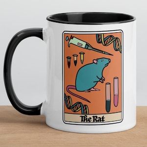 May include: White ceramic mug with a black rim and handle. The mug features a colorful illustration of a rat, a syringe, test tubes, and DNA strands. The text "The Rat" is printed below the illustration.