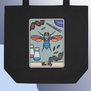 以下が含まれることがあります： 白いデザインの赤い、青い、そして黄色の羽を持つハエが描かれた黒いトートバッグ。ハエは試験管、ペトリ皿、DNAの鎖など、科学的なイメージに囲まれています。「The Fly」というテキストがハエの下に印刷されています。