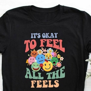 Puede incluir: Camiseta negra con el texto "It's okay to feel all the feels" en una fuente retro colorida. El texto está rodeado de un grupo de caras de emoji coloridas.
