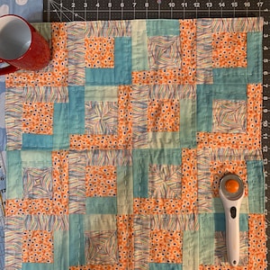 May include: A patchwork quilt with a variety of orange and blue patterned fabric squares. The quilt is being worked on with a rotary cutter and ruler.