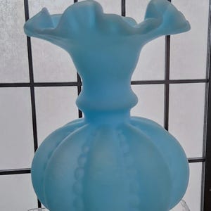 May include: A light blue glass vase with a ruffled top and a textured, bulbous body. The vase is sitting on a clear glass surface.