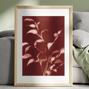 May include: A framed art print featuring a botanical design. The artwork showcases a close-up of plant leaves in shades of white and cream against a deep red background. The frame is made of light-colored wood, adding a natural touch to the piece.