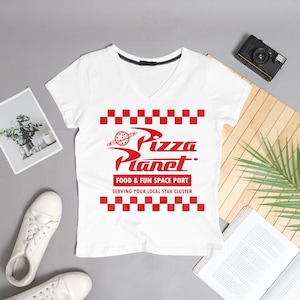 May include: White V-neck t-shirt with a red graphic of the Pizza Planet logo from the movie Toy Story. The logo features a red checkered pattern and the text "Pizza Planet. Food & Fun Space Port Serving Your Local Star Cluster".