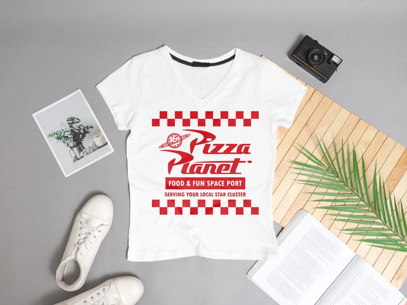 May include: White V-neck t-shirt with a red graphic of the Pizza Planet logo from the movie Toy Story. The logo features a red checkered pattern and the text "Pizza Planet. Food & Fun Space Port Serving Your Local Star Cluster".