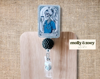 May include: A retractable badge reel featuring a rectangular design with a skeleton nurse holding a coffee cup. The image is in blue and white, with the text "THE NURSE PRACTITIONER". The reel has a black beaded top and a clear plastic clip.