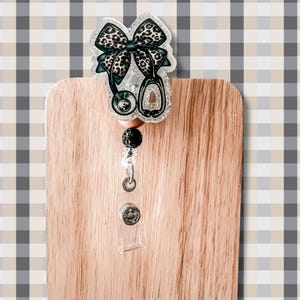Puede incluir: Un portatarjetas de acr&iacute;lico transparente con un dise&ntilde;o de estetoscopio y un lazo con estampado de leopardo. El portatarjetas tiene un cord&oacute;n retr&aacute;ctil, una cuenta negra y un clip de pl&aacute;stico transparente. El portatarjetas est&aacute; fijado a una superficie de madera.
